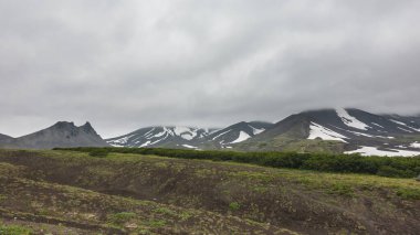 Dağlarda yoğun gri bulutlar uzanıyor. Yamaçlarda kar alanları var. Yerde seyrek bitki örtüsü var. Uzakta sisli bir gökyüzünün arka planına karşı resim gibi bir dağ. Kamçatka.