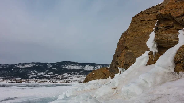 植生のない花崗岩の岩は 青空に対して 石の上に亀裂があり ベースはつららで覆われています 遠くの山の範囲と凍結湖 バイカル — ストック写真