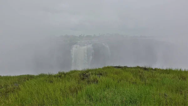 ビクトリアの滝の強力な流れは濃い霧の中に隠されています 手前は緑の芝生の牧草地 ジンバブエ — ストック写真