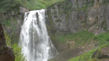 Şelale uçurumun kenarından aşağı akar. Etrafa su sıçrayan bir sis var. Rüzgar yeşil otları karıştırır. Kamçatka.