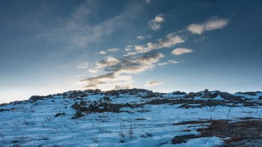Mavi gökyüzüne karşı karla kaplı bir tepe. Batan güneş bulutları altın bir ışıkla aydınlatır. Yerde kuru çimenler var. Sibirya