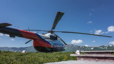 Helikopter sönmüş bir volkanın kalderasında yeşil bir çayırda duruyor. Mavi gökyüzüne karşı pitoresk bir dağ sırası. Ön planda ahşap bir merdiven var. Kamçatka.