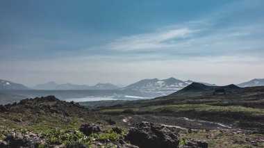 Dağ yamacında taramalı bitki örtüsü ve volkanik kayalar görülebilir. Uzakta, bir göl görebilirsiniz, mavi gökyüzüne karşı pitoresk bir dağ sırası. Kamçatka.
