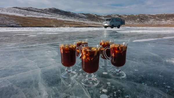 Four glass mugs with mulled wine are standing on a frozen lake. Hot red wine, sliced fruit. Cracks and bubbles of frozen methane are visible on the blue ice. There is a minibus on the shore. Baikal