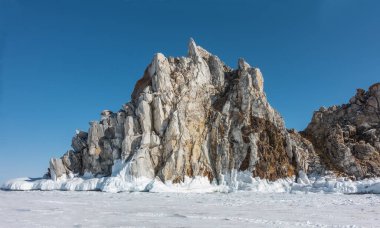A picturesque granite rock, devoid of vegetation, rise above a frozen lake. There are cracks on the stones. There is a layer of icicles at the base. Clear blue sky. Baikal  clipart