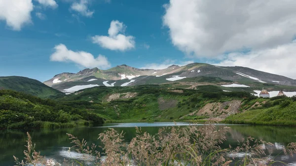 湖中的翡翠水很平静 蓝天和蓝云背景下的山脉 斜坡上有雪地 在草丛的前部 堪察加半岛 — 图库照片