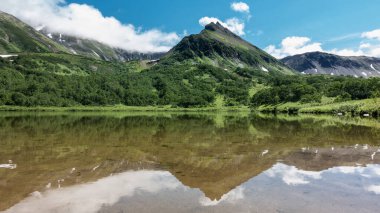 Gölün kıyısındaki pitoresk bir dağ sırası. Yamaçlarda yeşil bitki örtüsü ve erimiş kar var. Sakin suda bir ayna yansıması. Mavi gökyüzündeki bulutlar. Kamçatka. Vachkazhets