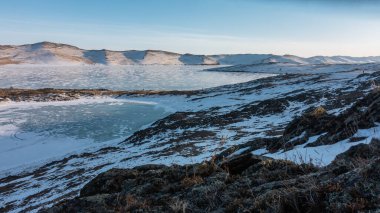İki donmuş göl ince bir toprak şeridiyle ayrılır. Kar hem buzda hem de yamaçlarda yatar. Ön planda kayalar ve kuru otlar vardır. Mavi gökyüzü. Güneşli bir kış günü. Baykal.