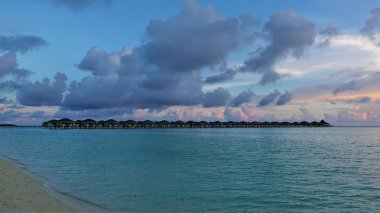 Bir sıra su villası akuamarin okyanusu üzerinde uzanır. Kumsalda küçük dalgalar dönüyor. Gökte mavi ve pembe bulutlar var. Maldivler