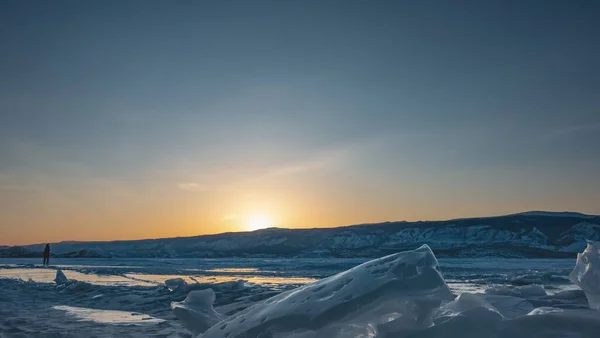 冬の夜 太陽は山の後ろに隠れており 空はオレンジ色で強調表示されています 凍った湖の上にグレア 氷の上に一人の男のシルエット 前景に ハンモックのブロックです バイカル — ストック写真