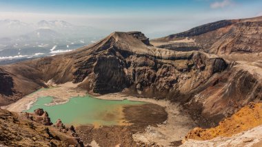 An amazing turquoise acid lake in the crater of a volcano. High steep slopes. There is snow on the banks, deposits of sulfur. In the distance, against the blue sky  a mountain range. Kamchatka. Gorely clipart