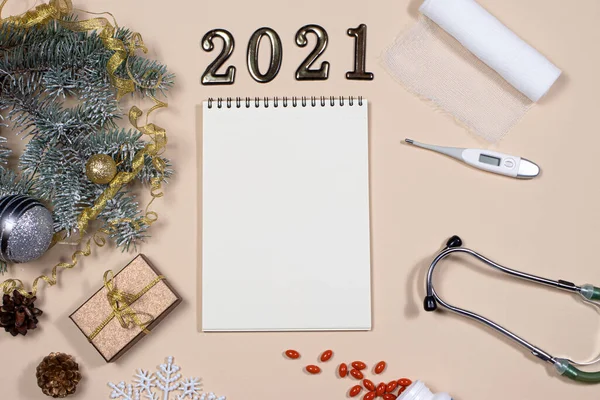 New Year\'s card on medical subjects: figures 2021, phonendoscope, tablets, thermometer and bandage with spruce branch on a beige background.