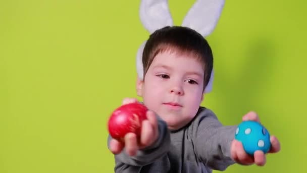 Alegre niño feliz con orejas de conejo en la cabeza está jugando con huevos de Pascua de colores, batiendo huevos de pollo pintados uno contra el otro. Niño de Pascua sobre un fondo amarillo. — Vídeo de stock