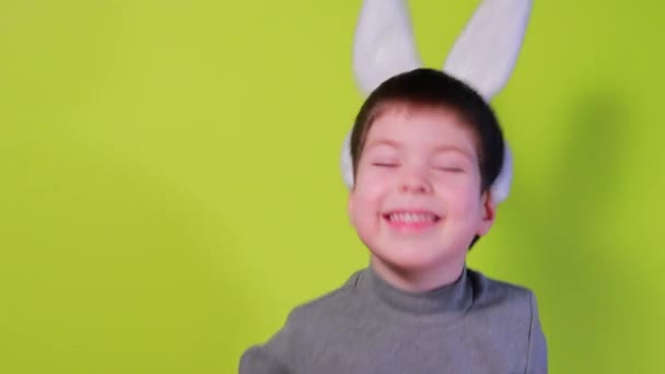 Счастливый смешной мальчик в кроличьих ушах на голове пытается сбросить уши с головы, кивает головой, играет и смеется на жёлтом фоне. Пасхальные дети — стоковое видео
