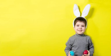 3-4 yaşlarında, gri ceketli, tavşan kulaklı neşeli bir çocuk. Renkli yumurtalarla oynuyor. Stüdyoda sarı bir arka planda paskalya yumurtası gösteriyor. Paskalya Bebeği