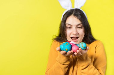 Kafasında tavşan kulakları olan mutlu bir esmer kadın paskalya yumurtaları tutuyor ve gülümsüyor. Paskalya için metin için kopyalama alanı olan sarı arkaplan üzerinde hazırlan