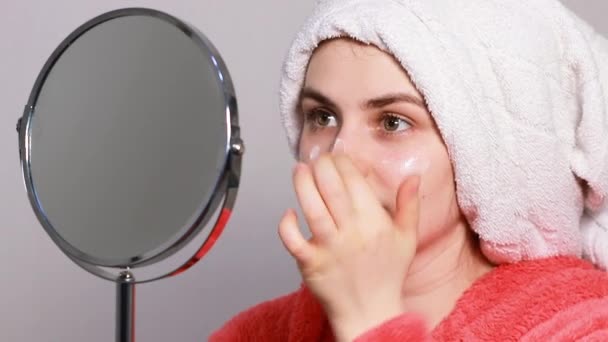 Młoda piękna kobieta z ręcznikiem na głowie nakłada krem na twarz i szyję siedząc przed lustrem. Naturalne kosmetyki do pielęgnacji skóry z witaminami i olejami, nawilżające i odżywcze. — Wideo stockowe