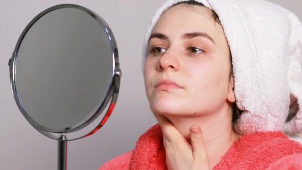 Μια νεαρή γυναίκα με ροζ μπουρνούζι κάνει μασάζ στο πρόσωπο, μια λευκή πετσέτα πέφτει από το κεφάλι της. Αστείο βίντεο για τη φροντίδα του δέρματος στην κοσμετολογία. — Αρχείο Βίντεο