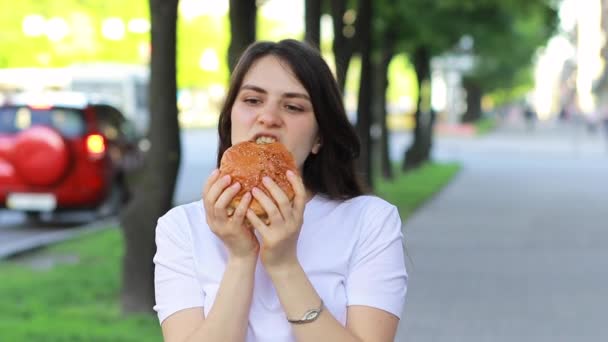 Brünette Frau isst Burger auf der Straße in der Nähe der Straße in der Stadt. Fast Food auf dem Weg zur Arbeit oder zur Schule. — Stockvideo