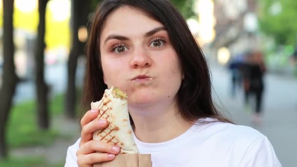 Mulher morena come shawarma em uma rua da cidade. Rua fast food pita roll com carne e legumes. — Vídeo de Stock