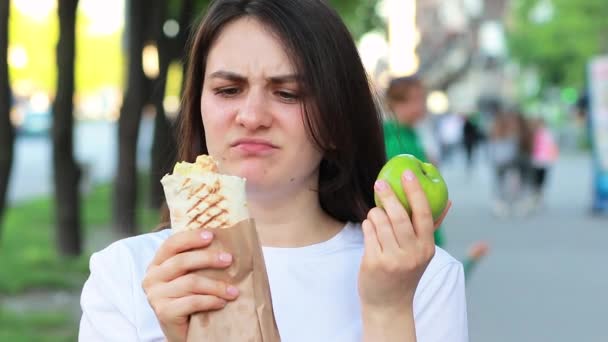 A bela mulher morena jovem em uma dieta escolhe a comida - que comer uma maçã ou shawarma de comida rápida. Morde a maçã e escolhe a dieta certa. — Vídeo de Stock