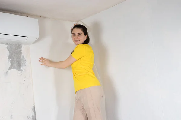 Une femme réparateur colle papier peint blanc sur le mur près du climatiseur sous le plafond. — Photo