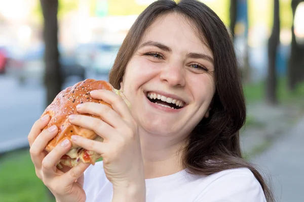 Mulher morena comendo hambúrguer e sorri na rua perto da estrada na cidade. Fast food a caminho do trabalho ou da escola. — Fotografia de Stock