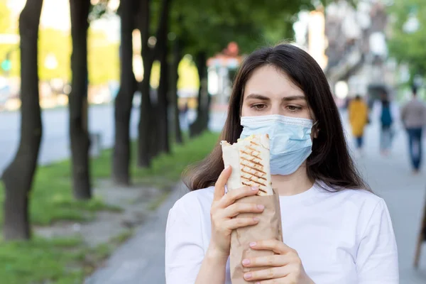 Una mujer enmascarada come shawarma de comida callejera. Comida rápida durante cuarentena y coronavirus, pandemia, cafés callejeros — Foto de Stock