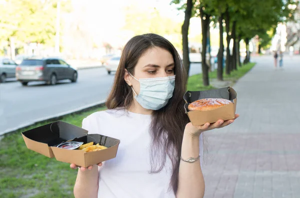 Una mujer enmascarada entregadora de alimentos durante una pandemia y cuarentena mantiene la comida a la orden en cajas de cartón. Comida callejera y coronavirus. — Foto de Stock