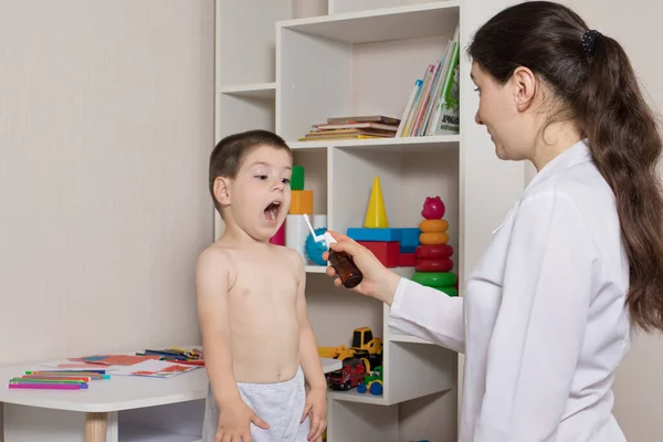 Egy gyermekorvos vagy nővér és egy 3-4 éves fiú. Torokspray, pharyngitis, stomatitis vagy szájpenész kezelése gyermekeknél. — Stock Fotó