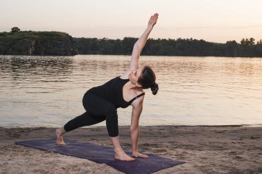 Gün batımında sahildeki bir paspasın üzerinde Yoga yapan kız ayakta duruyor. Genişletilmiş Kenar Açı Pozu Utthita Parsva Konasana