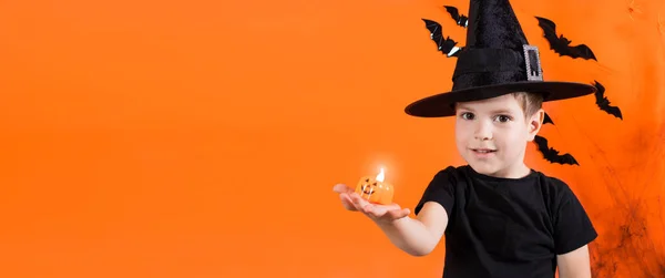 En pojke i en häxdräkt i svart hatt har en liten ficklampa som lyser pumpa Jack. på orange bakgrund. Halloween-ungar. Banner plats för text — Stockfoto