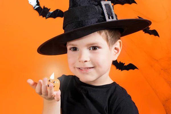 Un niño con un disfraz de bruja con un sombrero negro sostiene una pequeña linterna brillante lámpara de calabaza Jack. sobre un fondo naranja. Halloween niños. — Foto de Stock