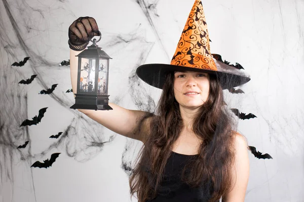 A woman in a witch costume in a hat and gloves in the background with bats holds a glowing lantern or lamp.. Halloween people.