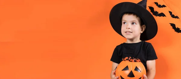 Halloween-barn. En förskolekille i häxdräkt har en pumpa med godis på orange bakgrund. Banner plats för text. — Stockfoto