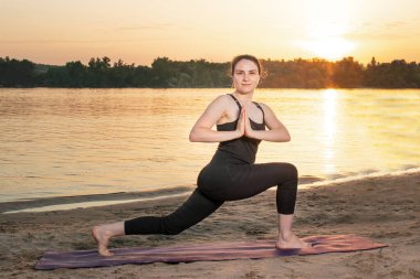 Bir kadın, Savaşçı 1 veya Virabhadrasana 1 pozisyonunda dururken yoga yapar. Yoga gün batımında doğada nehir kenarında.