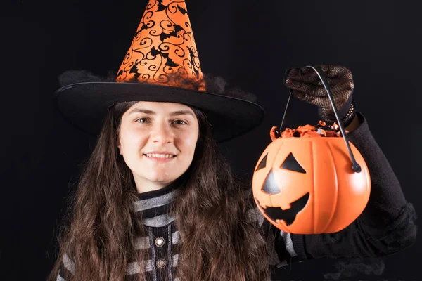 Una mujer con un traje de bruja en un sombrero y guantes sobre un fondo negro sostiene la calabaza cubo con dulces y sonrisa. Halloween personas —  Fotos de Stock