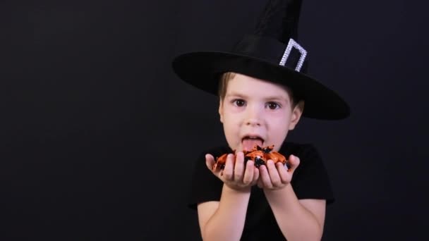 Halloween kind. Een kleuter in een heksenkostuum houdt een snoepje op een zwarte achtergrond, likt haar lippen en bereidt zich voor op de feestdag.. — Stockvideo