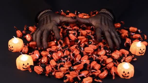 Una mujer en un disfraz de bruja rastrillos puñados de dulces con la inscripción Halloween sobre un fondo negro con velas de calabaza. Preparación para las vacaciones. — Vídeo de stock