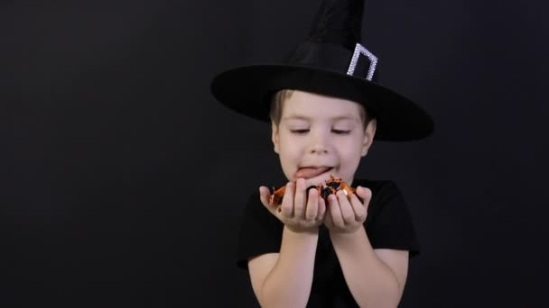 Halloweenkind. Een jongen in een heksenkostuum houdt een snoepje op een zwarte, likt haar lippen, snoep valt — Stockvideo