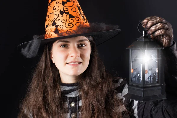 Uma mulher com um traje de bruxa em um chapéu e luvas em um fundo preto segura uma lanterna ou lâmpada brilhante. Pessoas de Halloween. — Fotografia de Stock