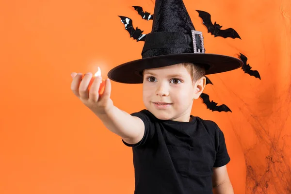 Un niño con un disfraz de bruja con un sombrero negro sostiene una pequeña linterna brillante lámpara de calabaza Jack. sobre un fondo naranja. Halloween niños. — Foto de Stock