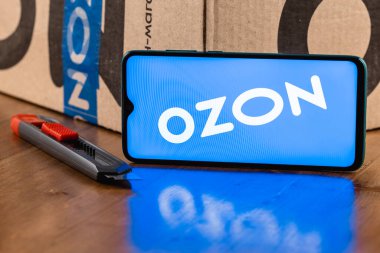 Ekran akıllı telefonunda Ozon logosu var. Yakın plan. Özon, Rusya 'daki ilk e-ticaret şirketlerinden biri. Paket ve maket kesicinin arka planında akıllı telefon var.