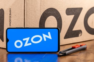 Ekran akıllı telefonunda Ozon logosu var. Yakın plan. Özon, Rusya 'daki ilk e-ticaret şirketlerinden biri. Paket ve maket kesicinin arka planında akıllı telefon var.