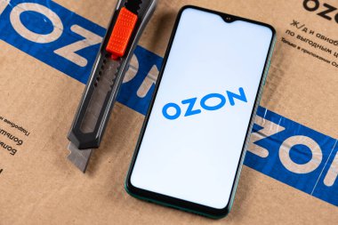 Ekran akıllı telefonunda Ozon logosu var. Yakın plan. Özon, Rusya 'daki ilk e-ticaret şirketlerinden biri. Paket ve maket kesicinin arka planında akıllı telefon var.