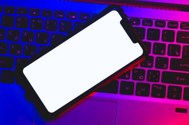 Laptop klavyesinde boş beyaz ekranlı bir akıllı telefonun neon ışıklandırmalı görüntüsü.