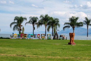 Jocotepec, Meksika - 22 Şubat 2025: Palmiye ağaçları ve Chapala Gölü yakınındaki çimenli bir parktaki heykel ile Jocotepec tabelasının görüntüsü