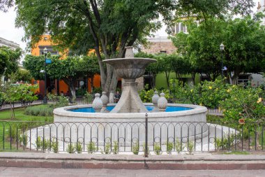 Tlaquepaque, Meksika - 5 Ekim 2025: Tlaquepaque, Meksika 'da canlı bir parkta mavi suyu olan geleneksel bir taş çeşmesi. Koloni mimarisi, yemyeşil ağaçlar ve canlı bir atmosfer.