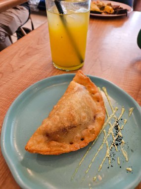 Yeşil soslu ve koyu baharatlı mavi bir tabakta altın renginde börek, tuzlu börek. Tahta bir masada serinletici soğuk bir içecek.