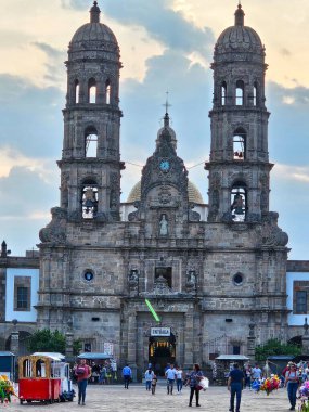 Meksika 'nın Jalisco kentindeki tarihi Zapopan Bazilikası' nın ayrıntılı ön görüntüsü. Özenle hazırlanmış Barok cephesi, ikiz çan kuleleri ve bulutlu gökyüzünün altında ziyaretçileri ve piyasa aktiviteleriyle birlikte kubbe.